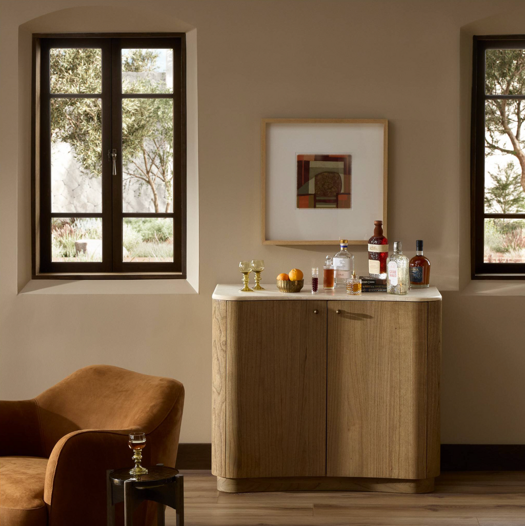 Wooden bar cabinet with drinks and decor in a room with windows and a chair.