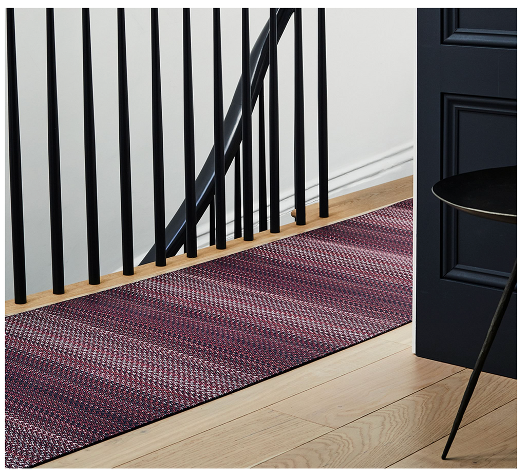 Purple braided rug on a wooden floor in front of a black door and staircase.