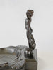Metallic sculpture of a nude figure on a decorative base against a white background