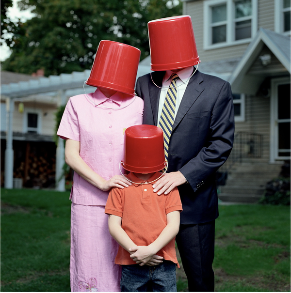 Three people with red buckets over their heads in front of a house.
