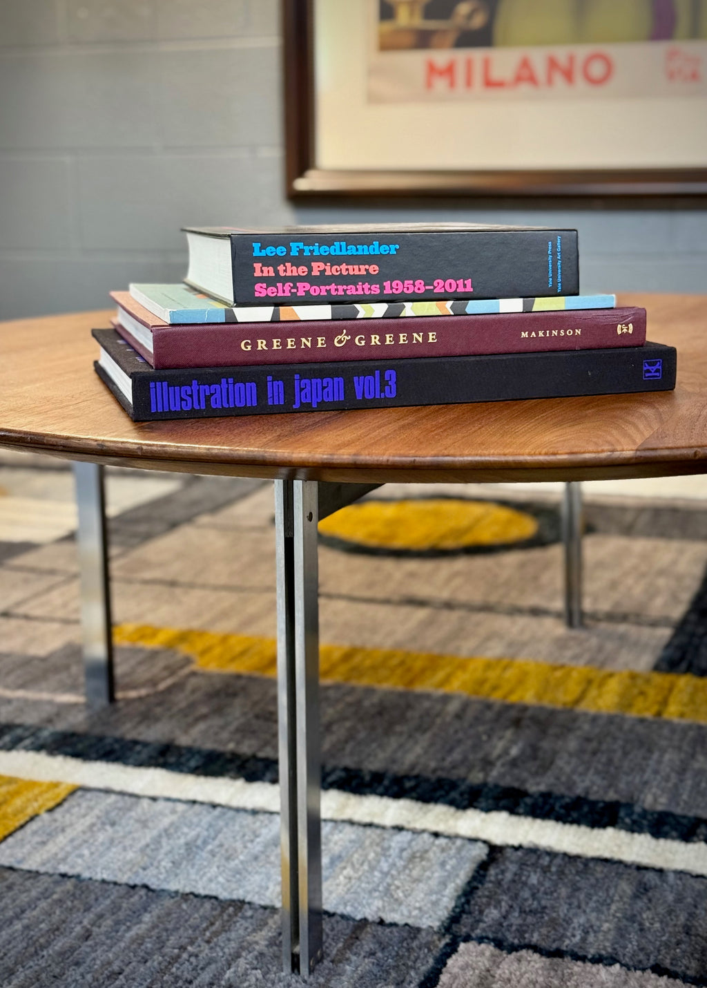 A stack of four books on a round wooden table in a modern room setting with a patterned rug and a framed print in the background. The books are titled 'Lee Friedlander In the Picture Self-Portrait 1958-2011', 'Greene & Greene Maximals', and 'Illustration in Japan vol.