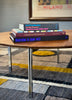 A stack of four books on a round wooden table in a modern room setting with a patterned rug and a framed print in the background. The books are titled 'Lee Friedlander In the Picture Self-Portrait 1958-2011', 'Greene & Greene Maximals', and 'Illustration in Japan vol.