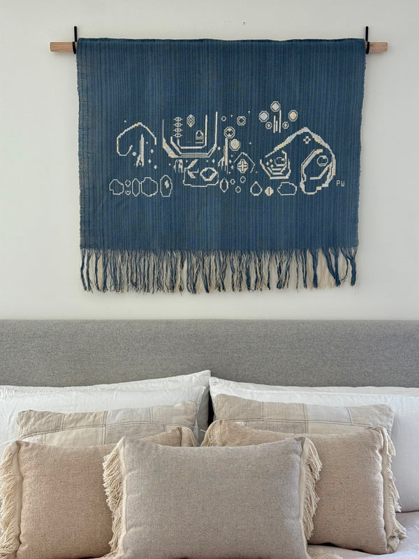 Blue woven tapestry with white patterns hanging above a sofa with beige pillows.