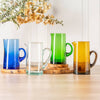 Four colorful glass mugs on a wooden surface with a vase of flowers in the background.