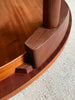 Close-up detail of a dark brown wooden table corner with a curved apron, leg joint, and visible brand stamp reading 'JENNY BURNS' on a light beige carpet.