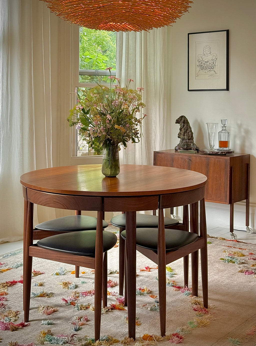 A round wooden dining table with four matching chairs featuring black upholstered seats, styled in a bright, modern dining room with a large woven pendant light, floral centerpiece, and a sideboard in the background.
