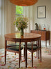 A round wooden dining table with four matching chairs featuring black upholstered seats, styled in a bright, modern dining room with a large woven pendant light, floral centerpiece, and a sideboard in the background.