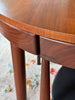 Close-up of a round wooden table with a warm medium-brown finish, showing the tabletop edge, drawer front, legs, and a small dark line detail on the drawer apron.