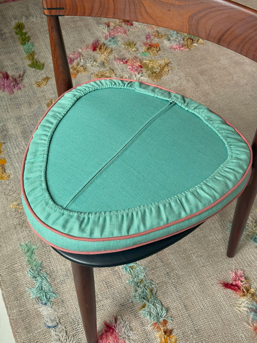 A close-up of a round upholstered cushion or seat pad in a muted sage green fabric with a pinkish-red piped edge, shown resting on a wooden chair over a textured beige rug.