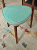 A mid-century modern stool with a rounded, asymmetrical seat cushion upholstered in a textured mint green fabric, finished with a contrasting pink piping trim and dark brown wooden legs, set on a textured beige rug.