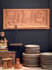 Wooden cutting board with carvings of kitchen utensils against a dark wall, with stacks of ceramic plates and wooden stools in the foreground.